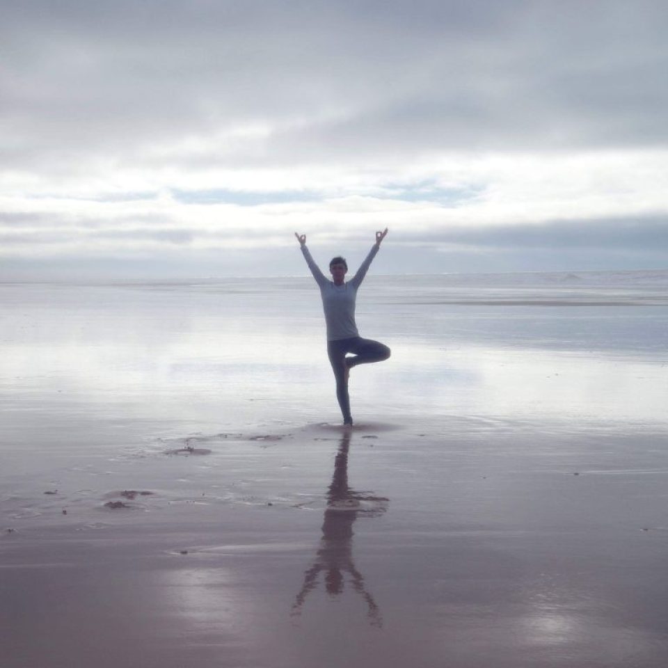 femme debout une jambe replié et les deux bras tendu vers le ciel, en position de yoga à la plage