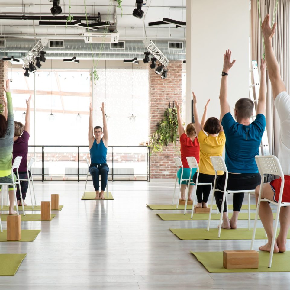 group of senior people doing yoga with chairs and with attractive young male teacher indoor