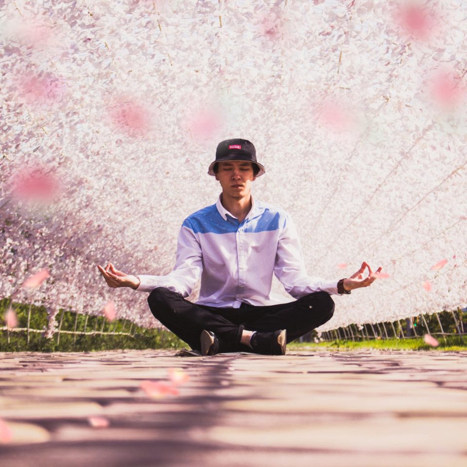 A young man in a hat meditates in a vibrant outdoor floral tunnel, embodying serenity and fashion.