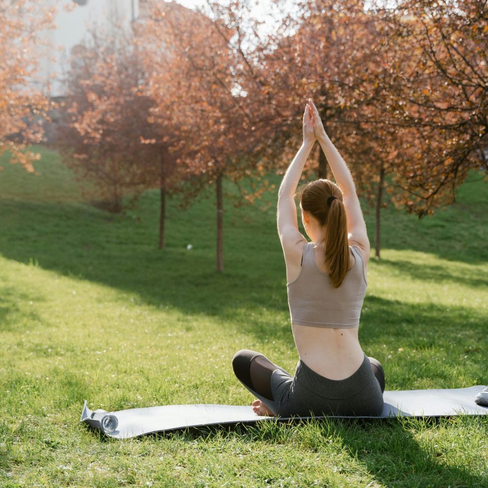 A woman practicing yoga on a mat outdoors in a sunlit park during autumn.