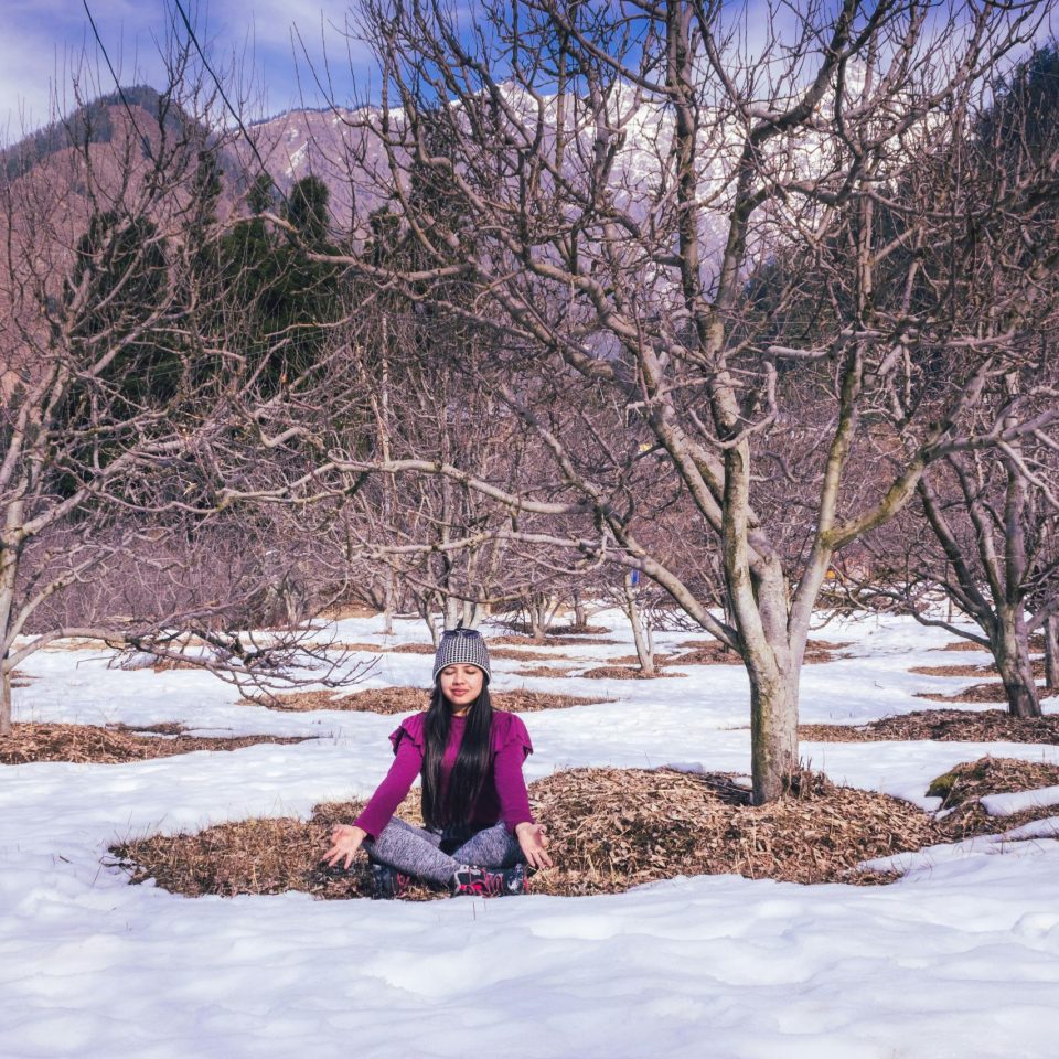 A woman meditates in lotus pose amidst snow-covered bare trees in a serene winter orchard.
